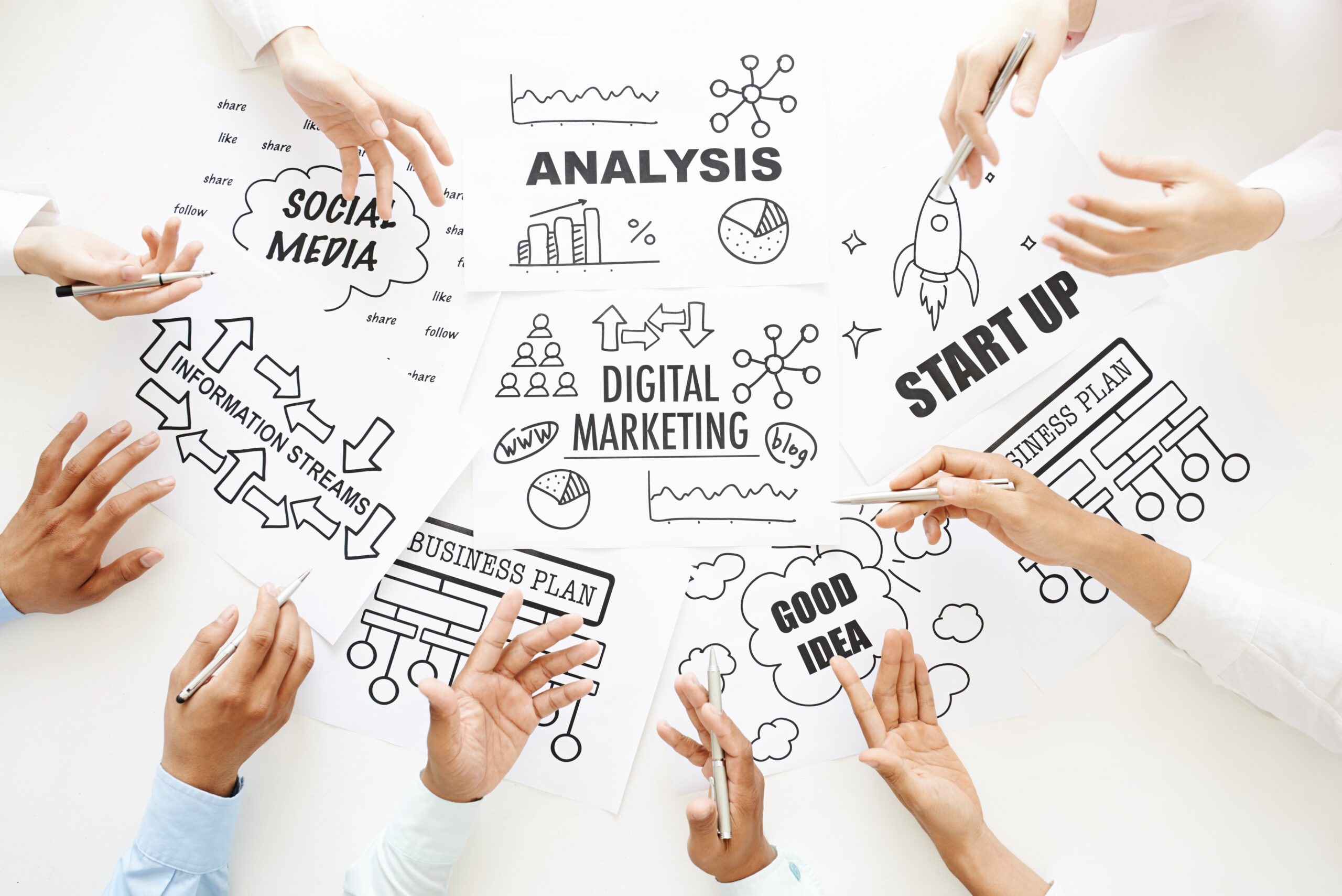 A top-down view of a marketing team’s hands pointing to sketches of business plans, social media icons, and digital marketing diagrams on a white table.