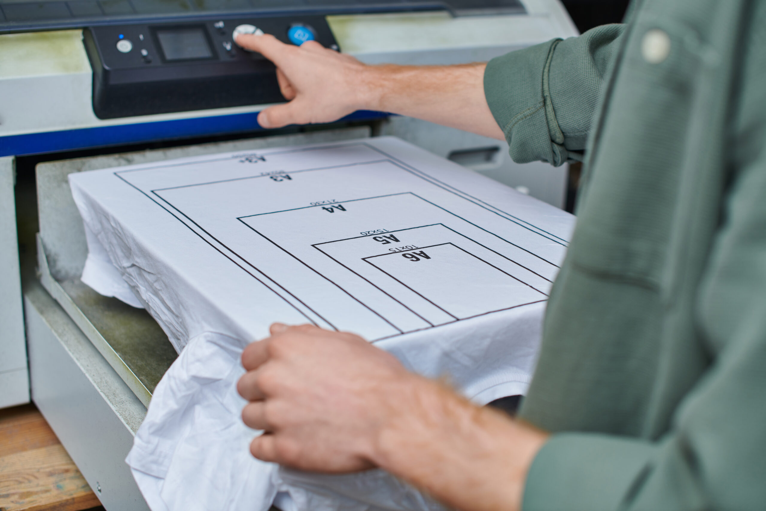 A worker operating a heat press machine to print paper size guides onto a white t-shirt, illustrating custom t shirt printing in Miami, FL.