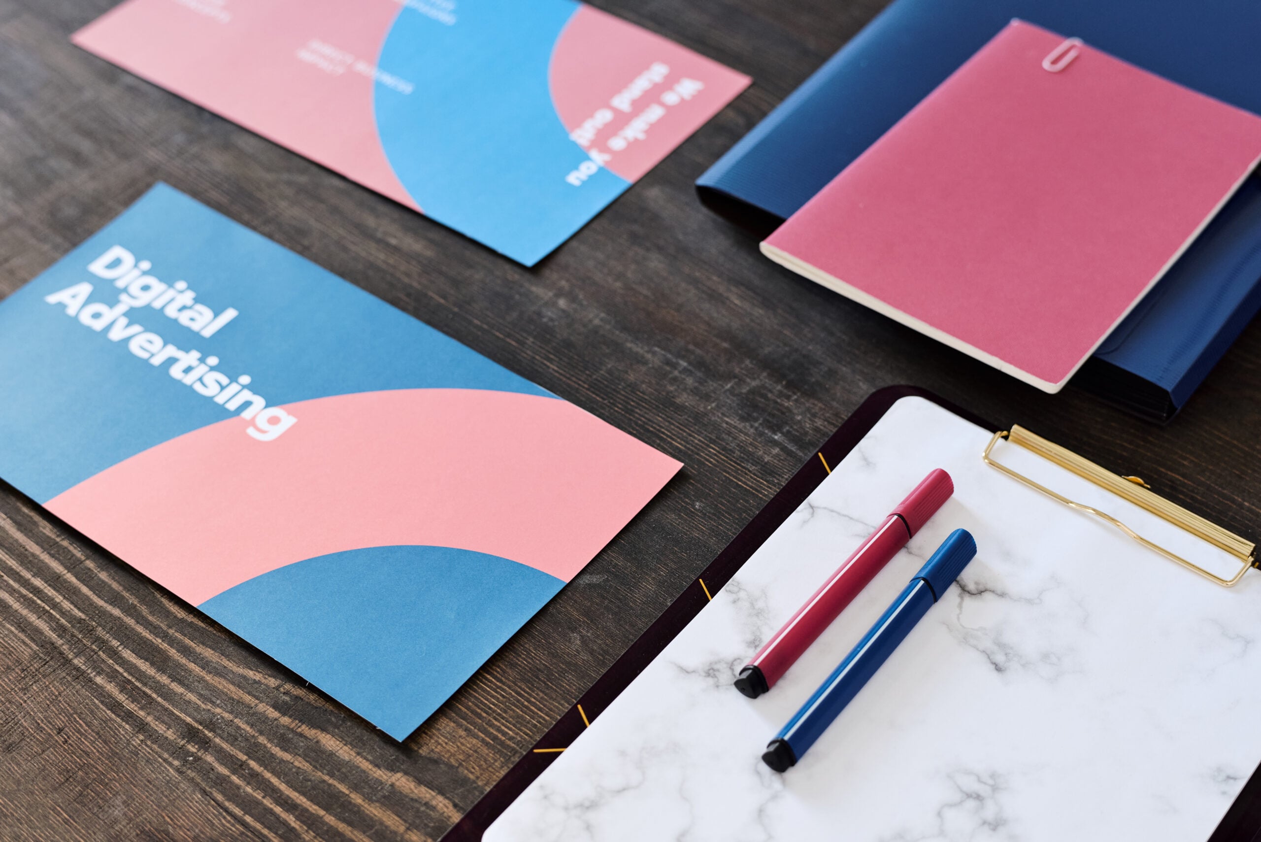 A top-down view of printed "Digital Advertising" brochures with blue and pink geometric designs, a marble-patterned clipboard, and colorful pens on a dark wooden desk.