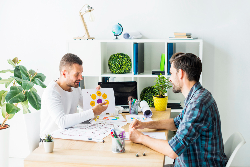 Two men collaborating at a bright wooden desk with blueprints and color charts while discussing interior design services in Denver, CO.