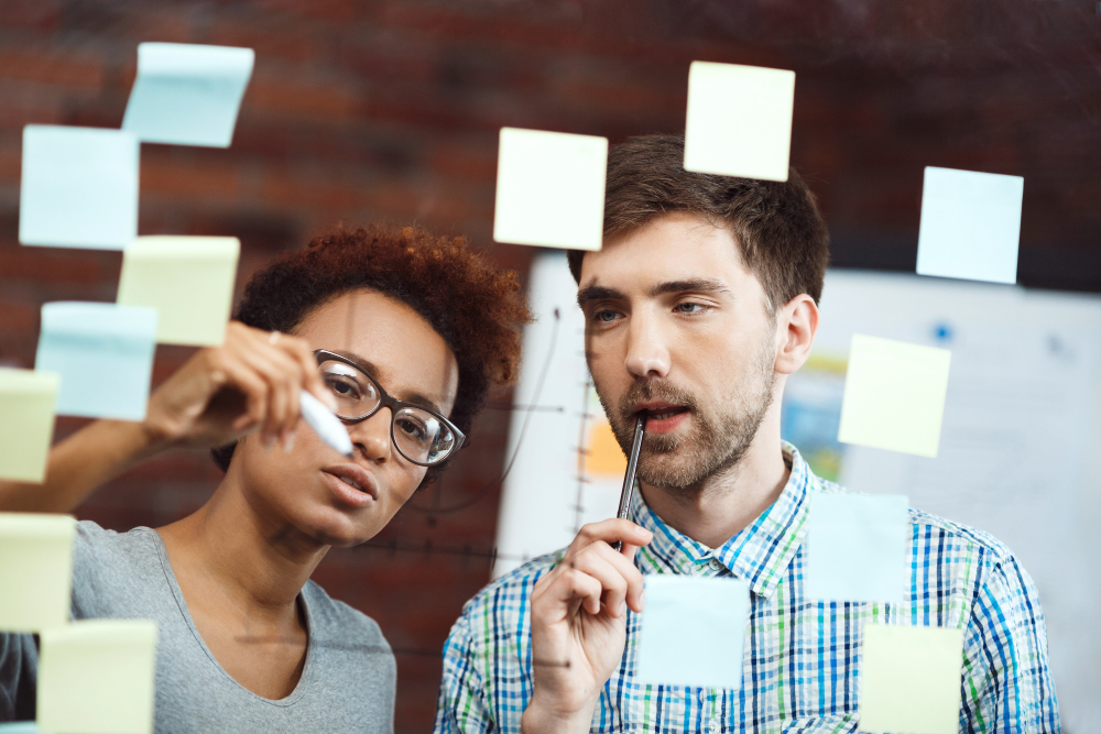 Two brand strategy consultants brainstorming ideas using sticky notes on a glass board in Newark, NJ.
