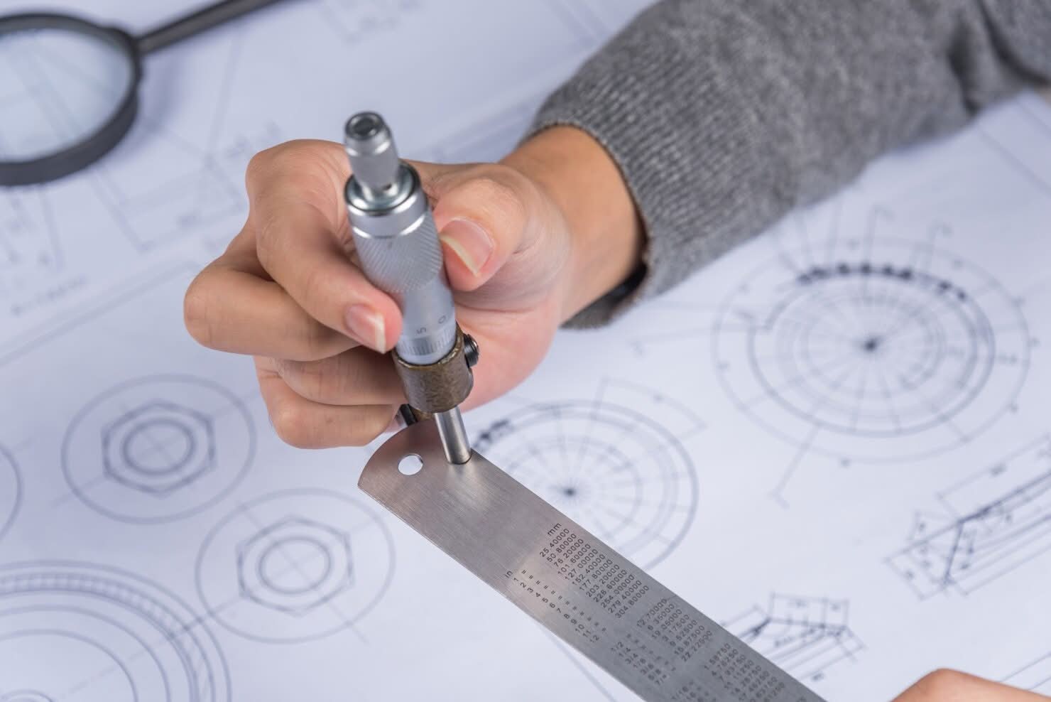 Engineer using a micrometer on a metal ruler over technical blueprints on a desk in Newark, NJ.