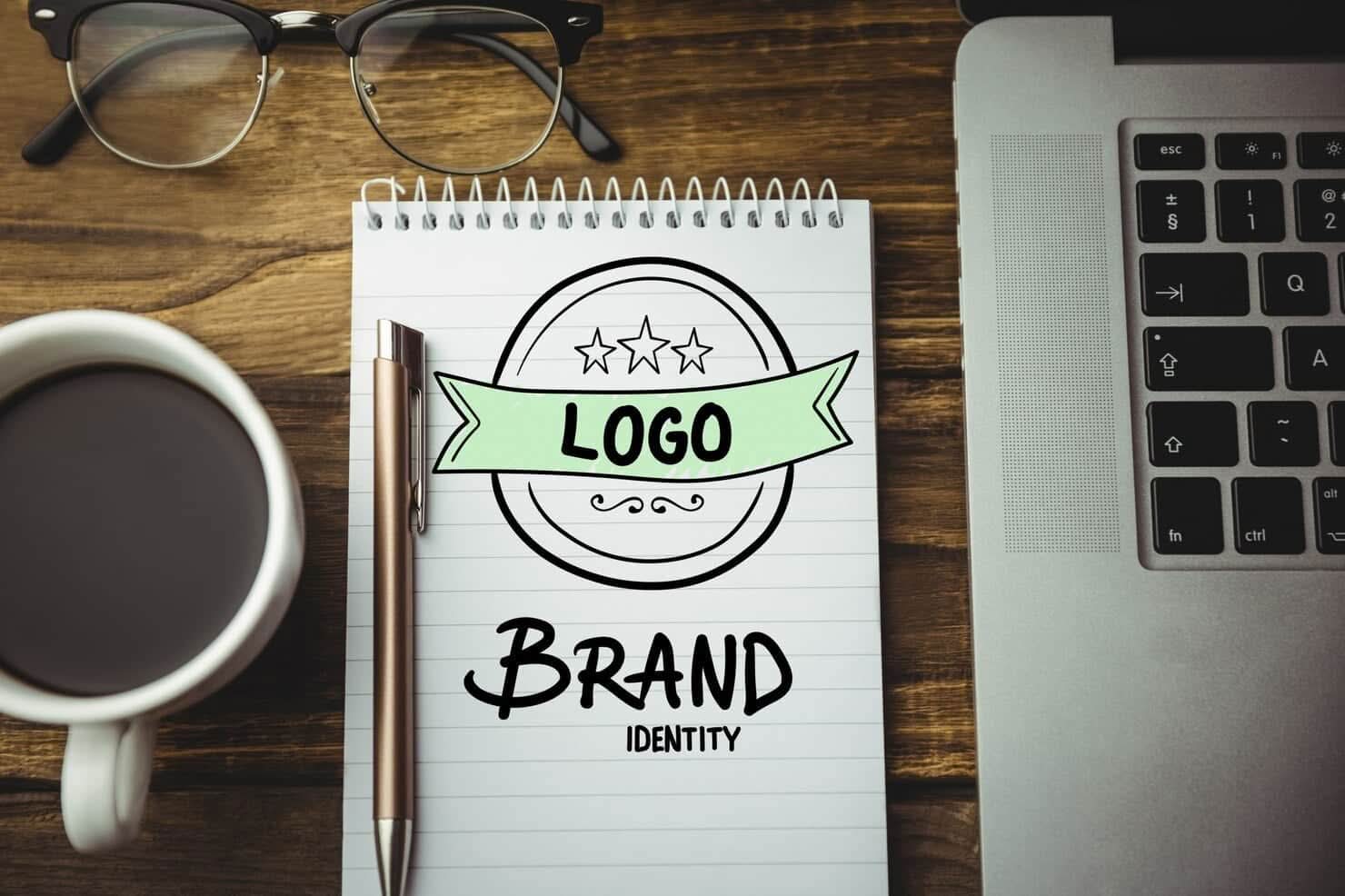 A top-down view of a wooden desk featuring a laptop, a cup of coffee, eyeglasses, and a notepad with a hand-drawn logo design and the words "Brand Identity."