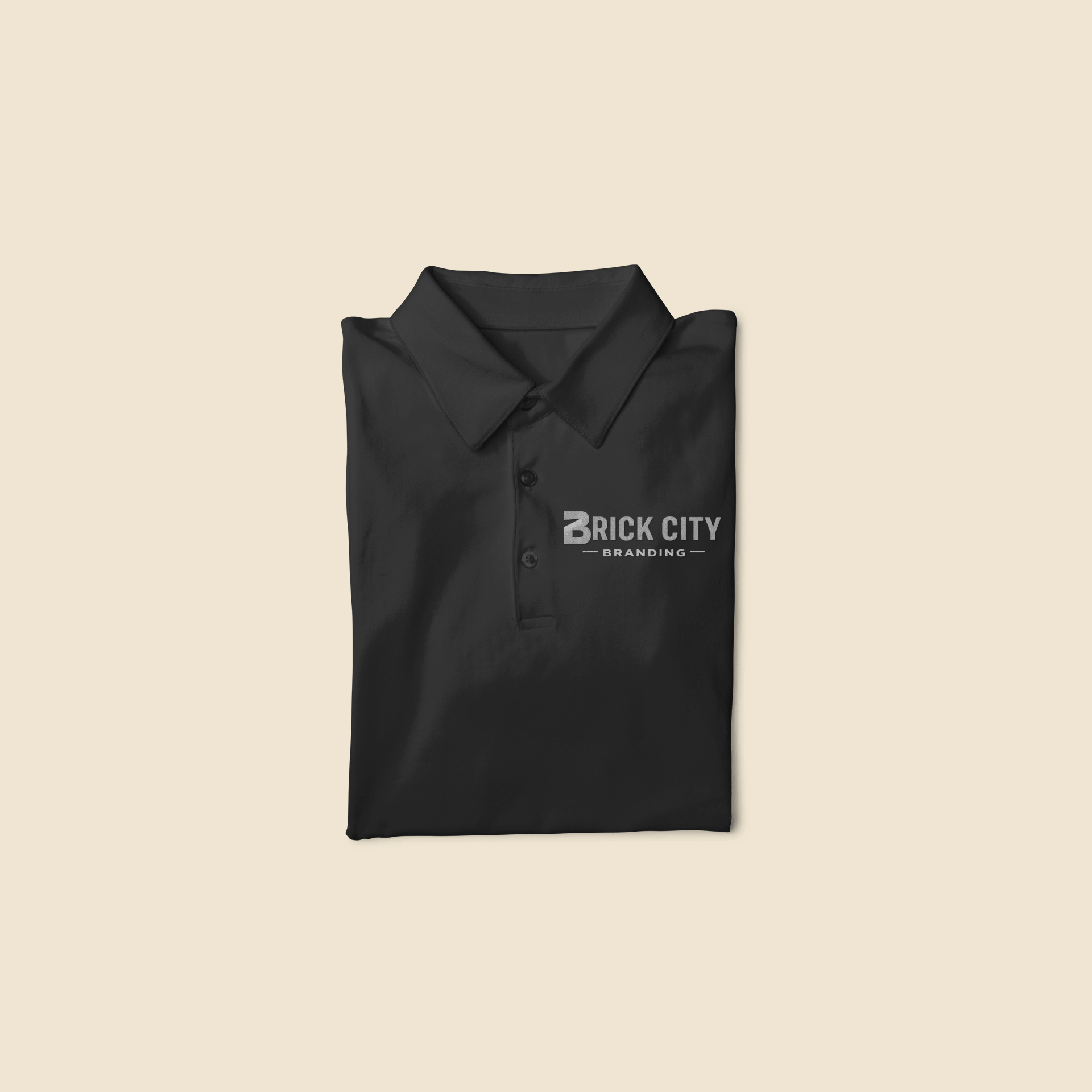 A neatly folded black branded polos shirt featuring a single-color silver or grey embroidered "Brick City Branding" logo on the left chest.