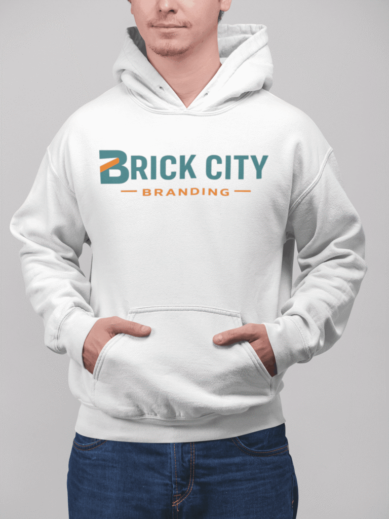A man with a beard and glasses wearing a white knit beanie with the Brick City Branding logo, posing in front of a rustic wooden barn or cabin.
