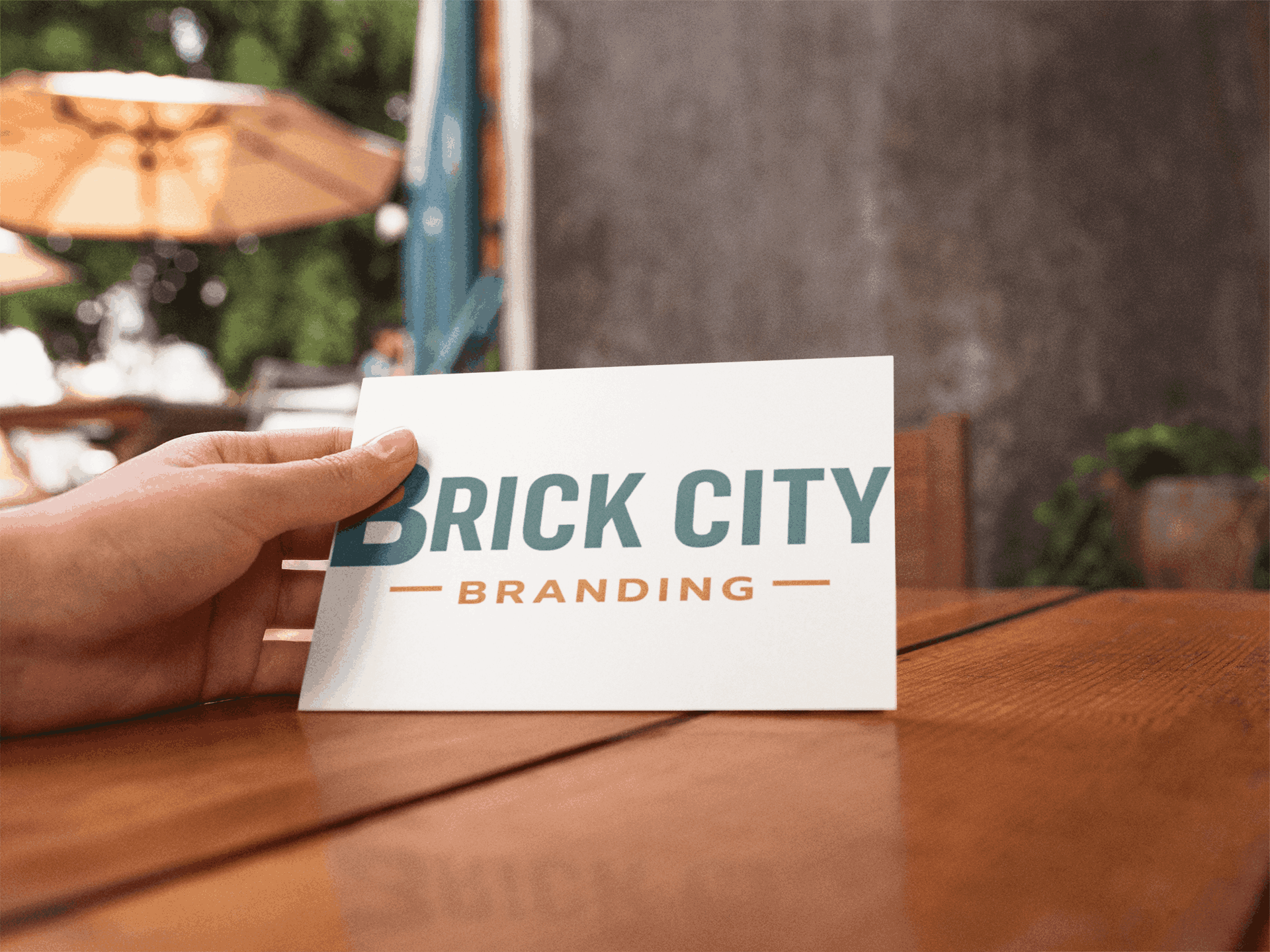A person's hand holding a white postcard with the "Brick City Branding" logo on a polished wooden cafe table with a blurred outdoor patio background.