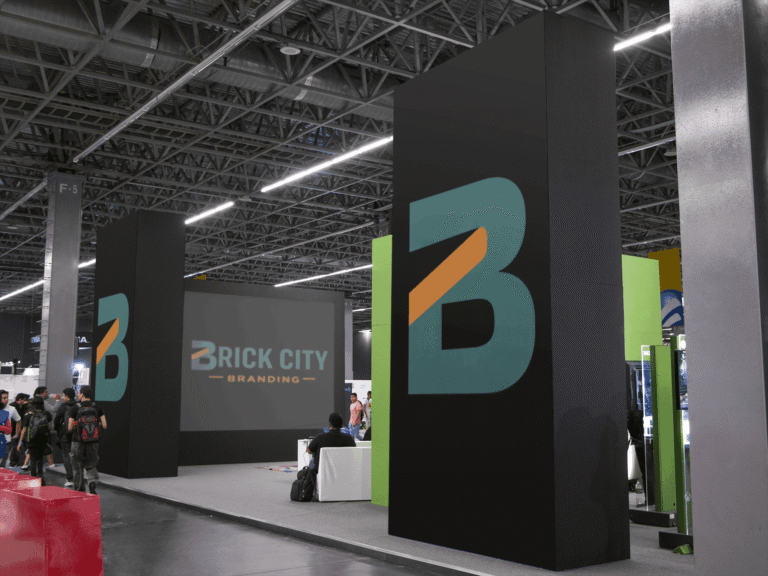 A large modern trade show exhibit and event branding for Brick City Branding featuring tall black monolith pillars with teal and orange logos and a large digital screen displaying the full brand name.