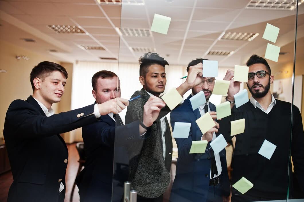 Logo Design 5 A group of five professional marketing strategists in business attire placing post-it notes on a glass wall during a brand development workshop.