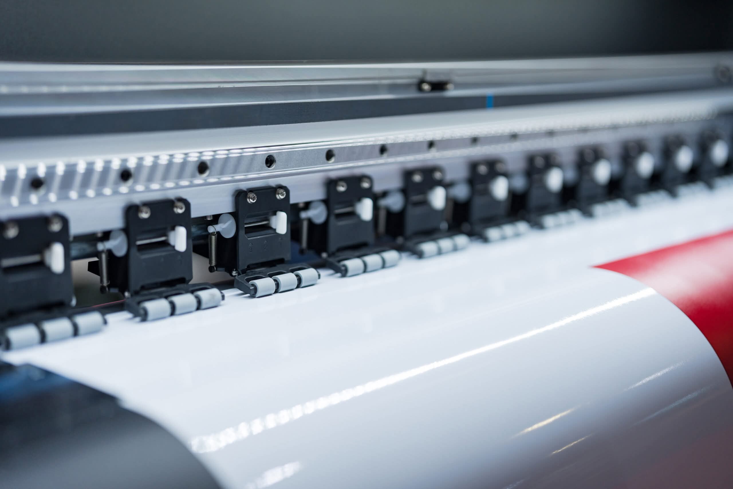 A close-up of a high-speed industrial wide-format printer feeding a roll of white vinyl with red graphic elements through precision rollers.