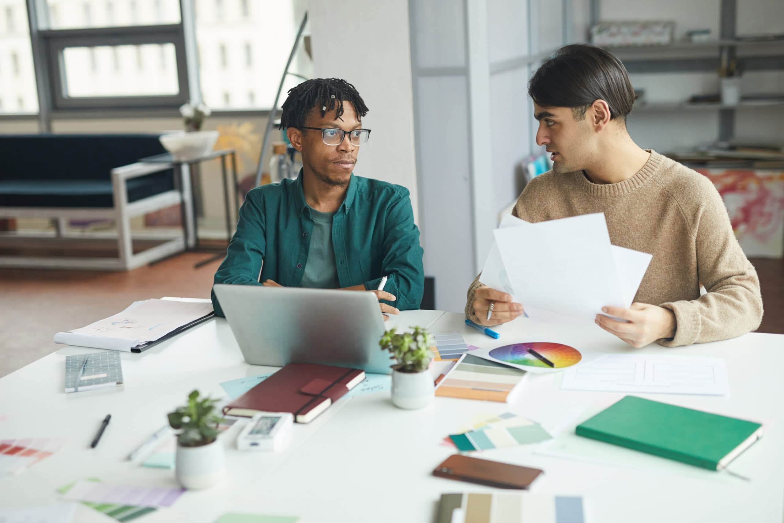 Two young professional designers collaborating over brand identity sketches and digital mockups in a bright, industrial-style office.