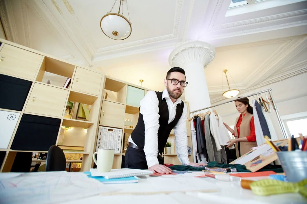 A male fashion designer wearing glasses leaning over a work table with sketches and fabric samples while an assistant checks garments, highlighting custom fashion designer services in New York, NY.