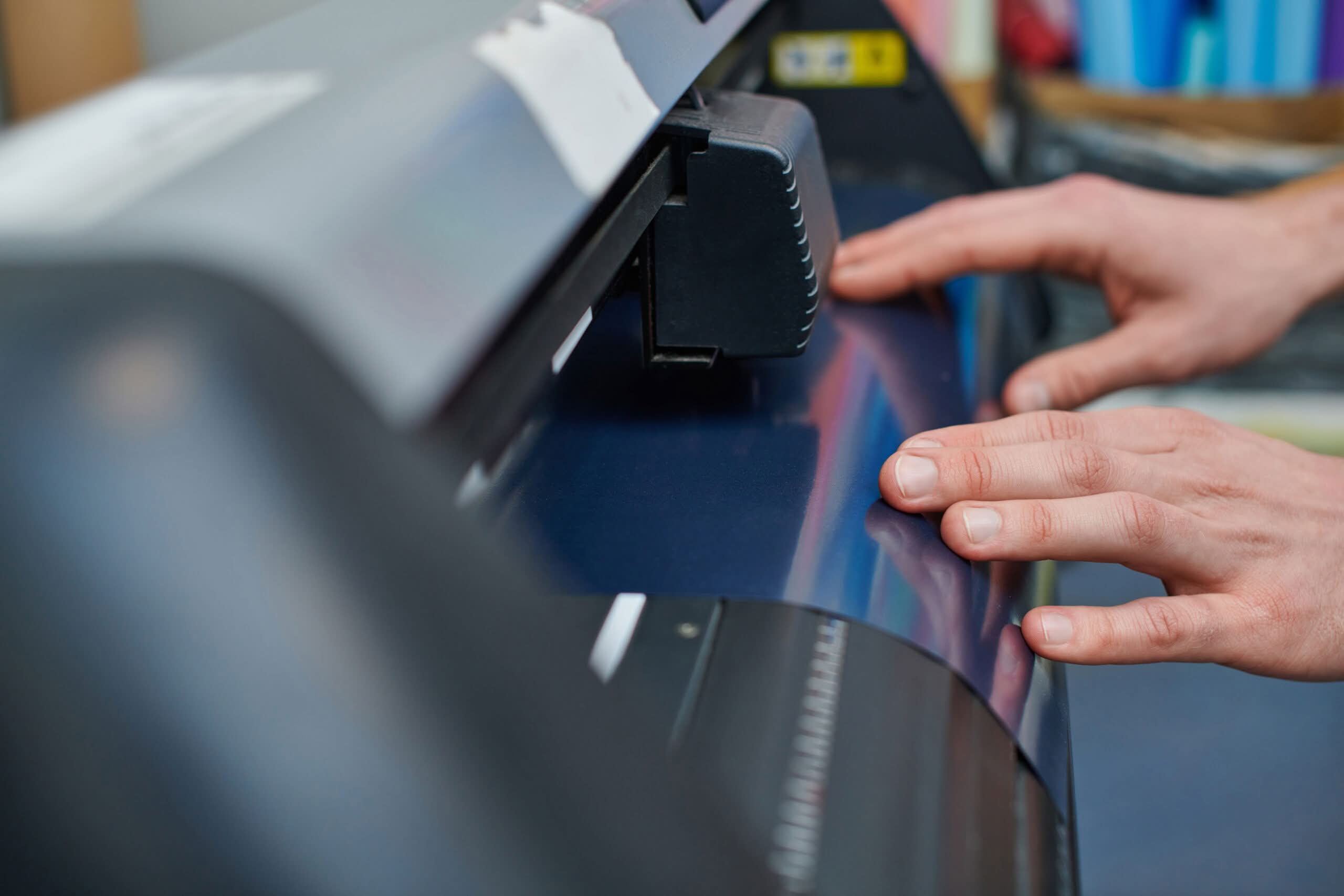 A close-up of a person's hands guiding blue vinyl signs through a professional cutting plotter machine in a production studio.