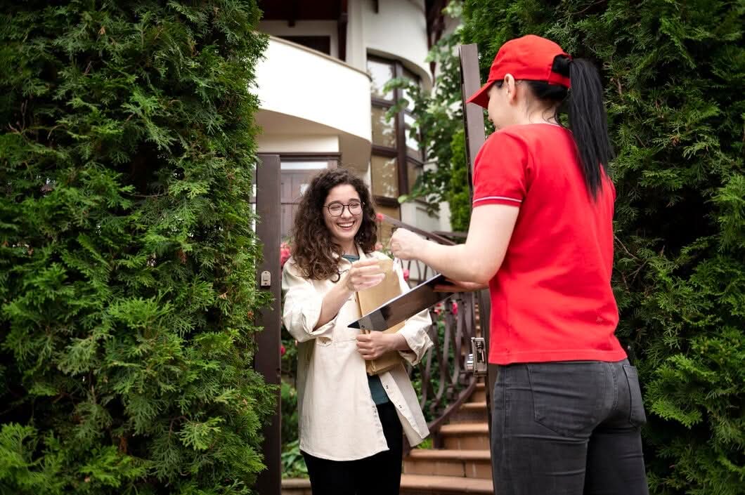 A friendly courier in a red uniform handing over a package and a clipboard for signature to a smiling customer at her doorstep.