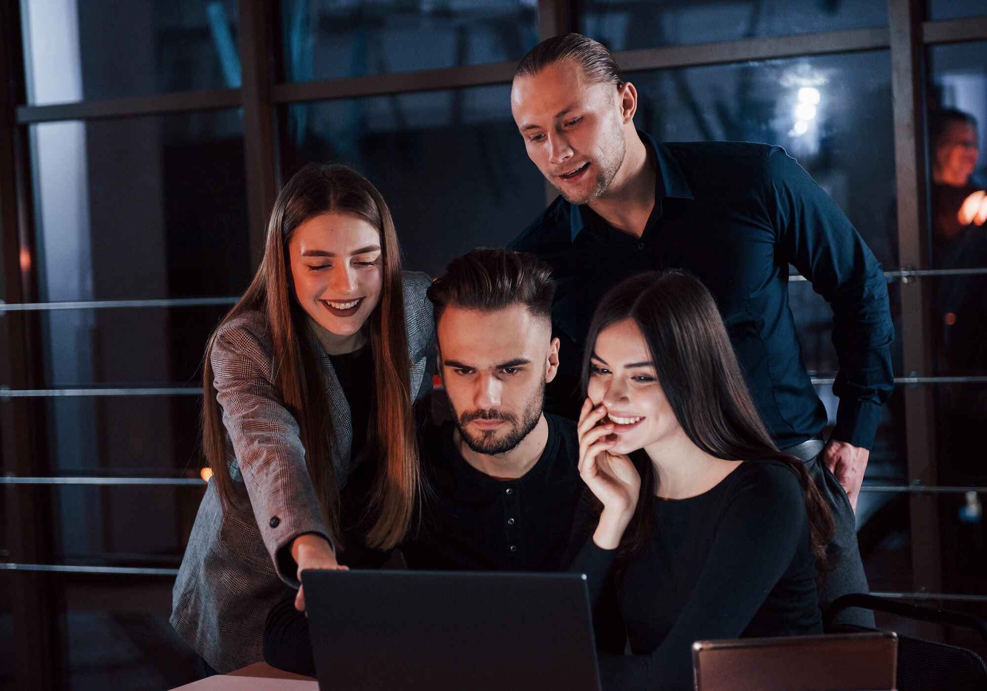 Contact a diverse group of four young business professionals in smart-casual attire smiling and collaborating in a bright, modern office with a city view.