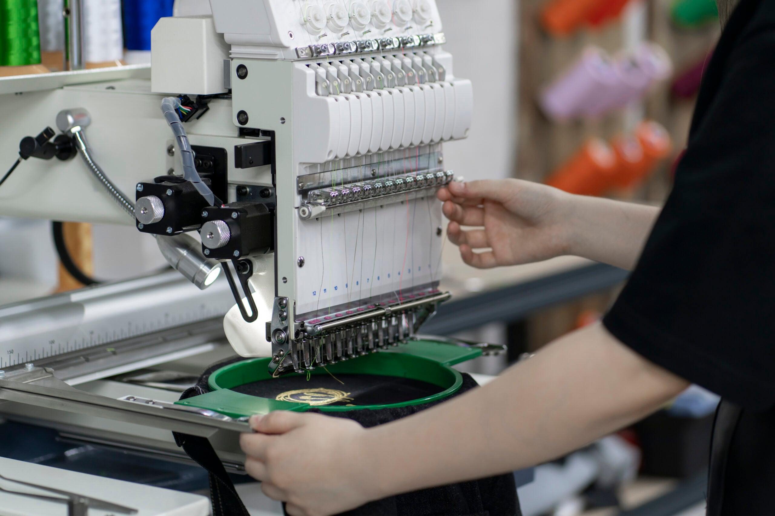 A worker aligning a green embroidery hoop containing a black cap under an industrial multi-needle machine, representing custom hat embroidery in Columbus, OH.