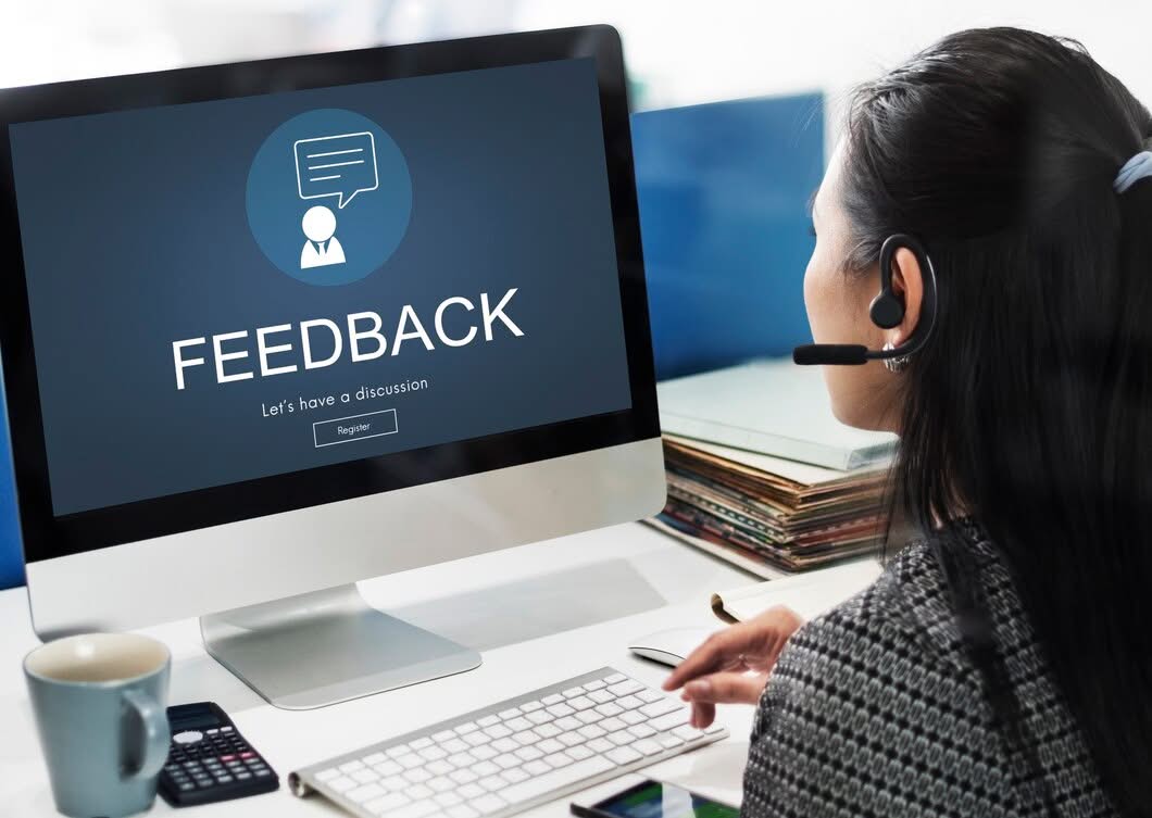 A female customer support representative wearing a headset, looking at a computer monitor displaying a "FEEDBACK - Let's have a discussion" registration screen.