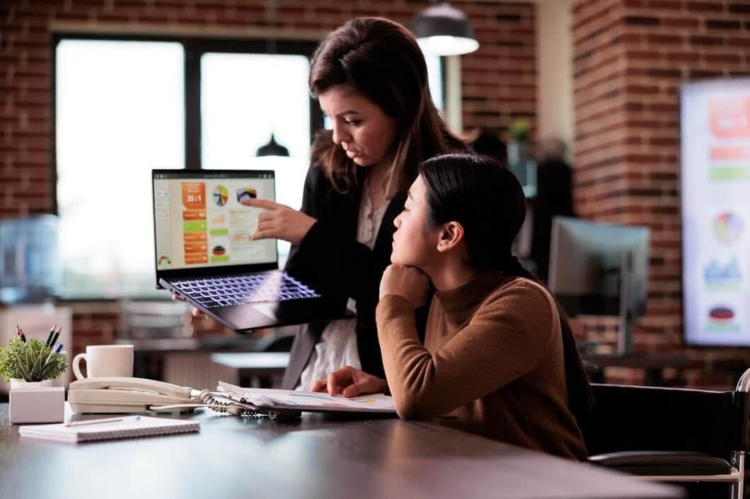 A marketing consultant pointing to colorful data charts and analytics on a laptop screen during a strategy session with a client in a modern brick-walled office.