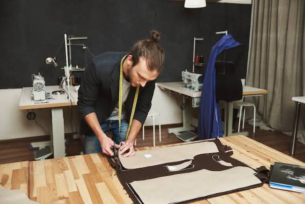 A male tailor leaning over a wooden table cutting fabric patterns with scissors, demonstrating custom tailor services in San Francisco, CA.