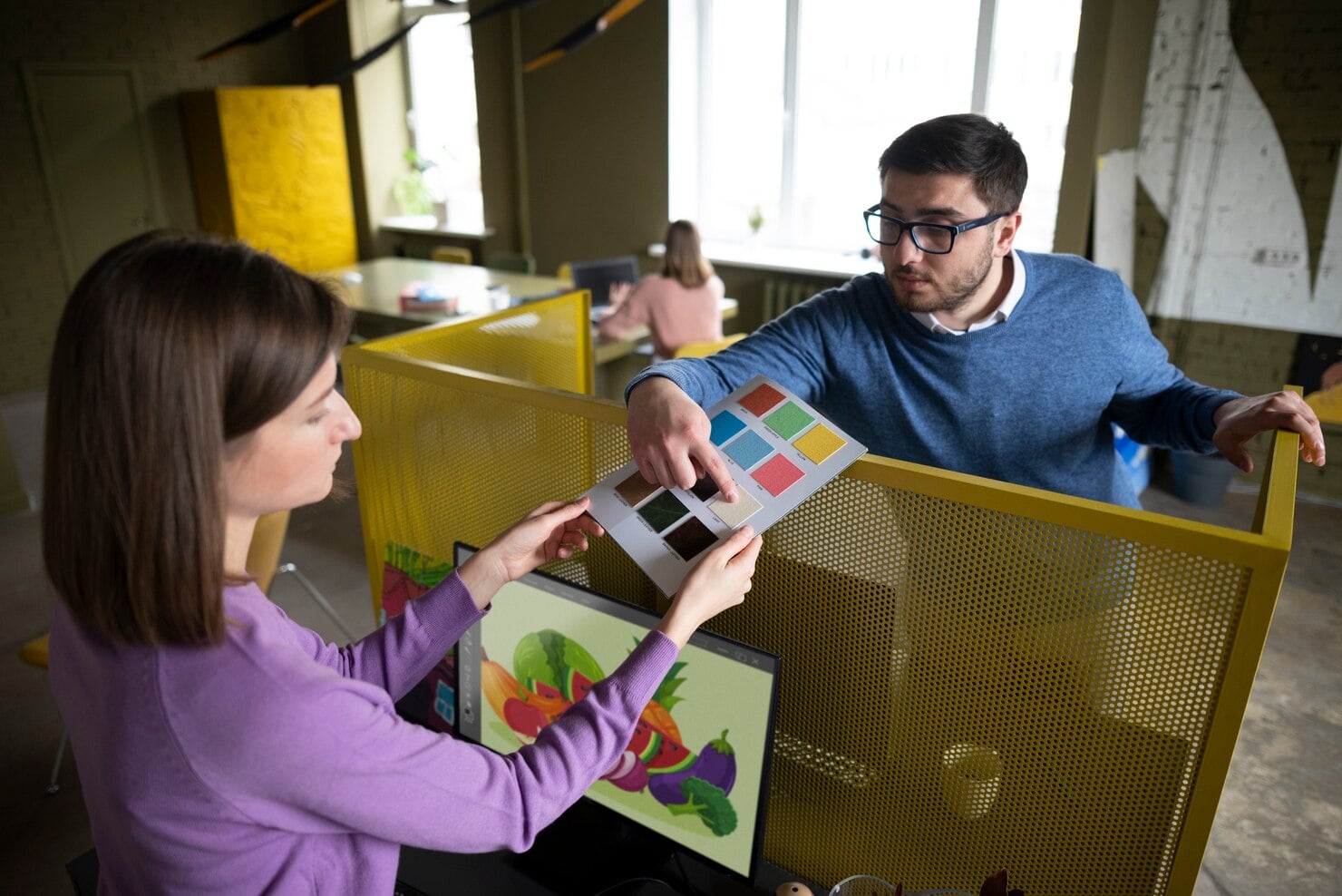 Two colleagues in a modern office with yellow partitions discussing a color palette while looking at a monitor displaying a vibrant vegetable illustration in a print services on Ocala.