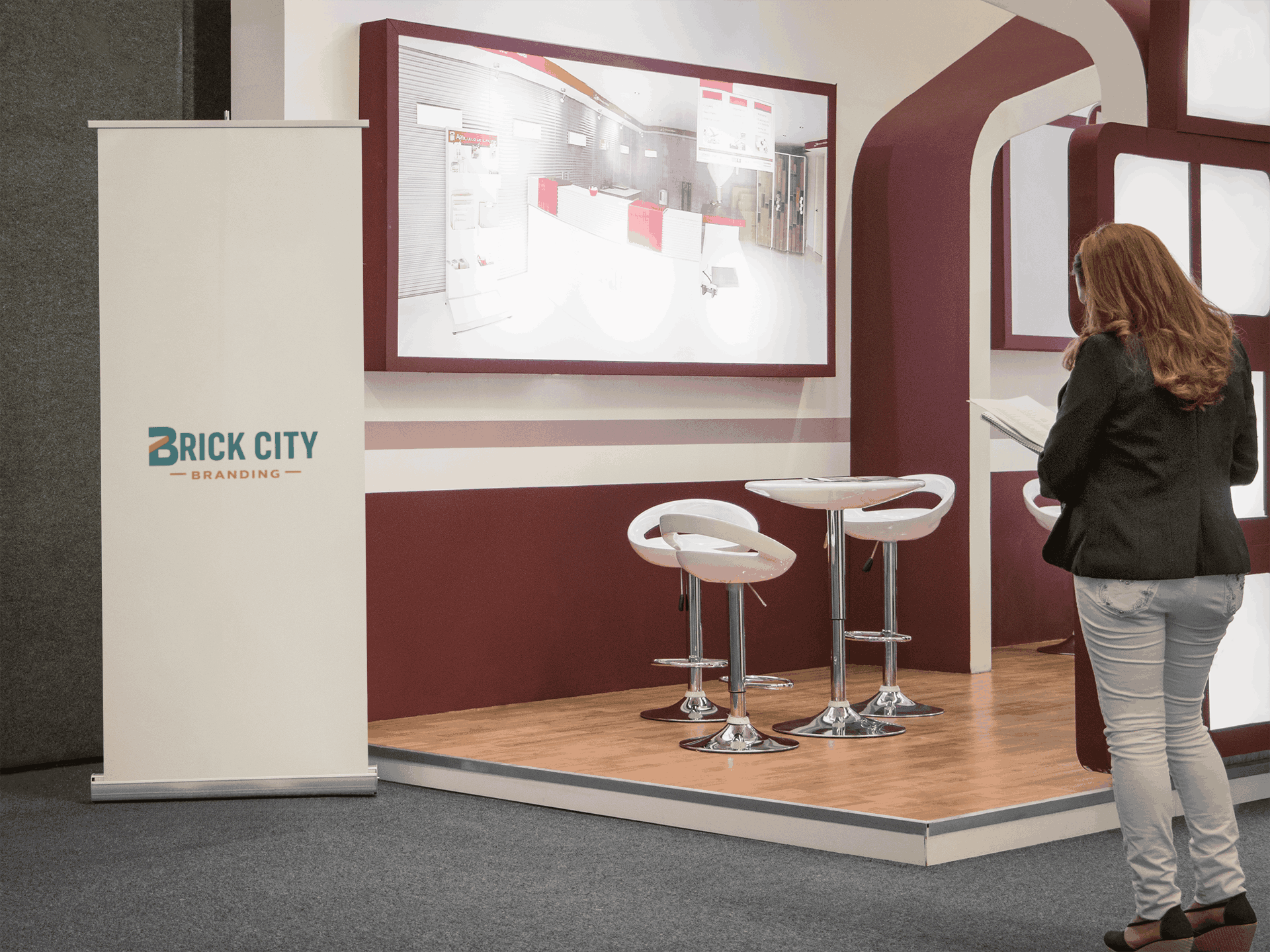 A vertical white retractable pull-up banner with the Brick City Branding logo positioned in a modern trade show booths with maroon accents and a white cafe table.