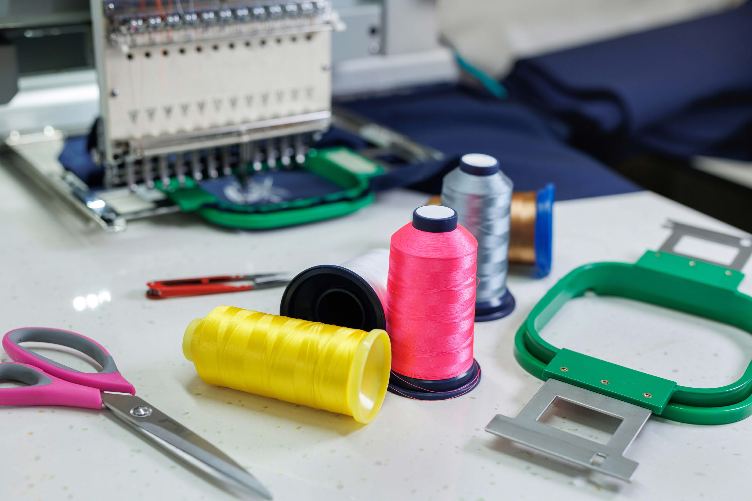 Colorful thread spools, scissors, and green embroidery hoops placed next to an industrial embroidery machine, showcasing custom embroidery services in Charlotte, NC.