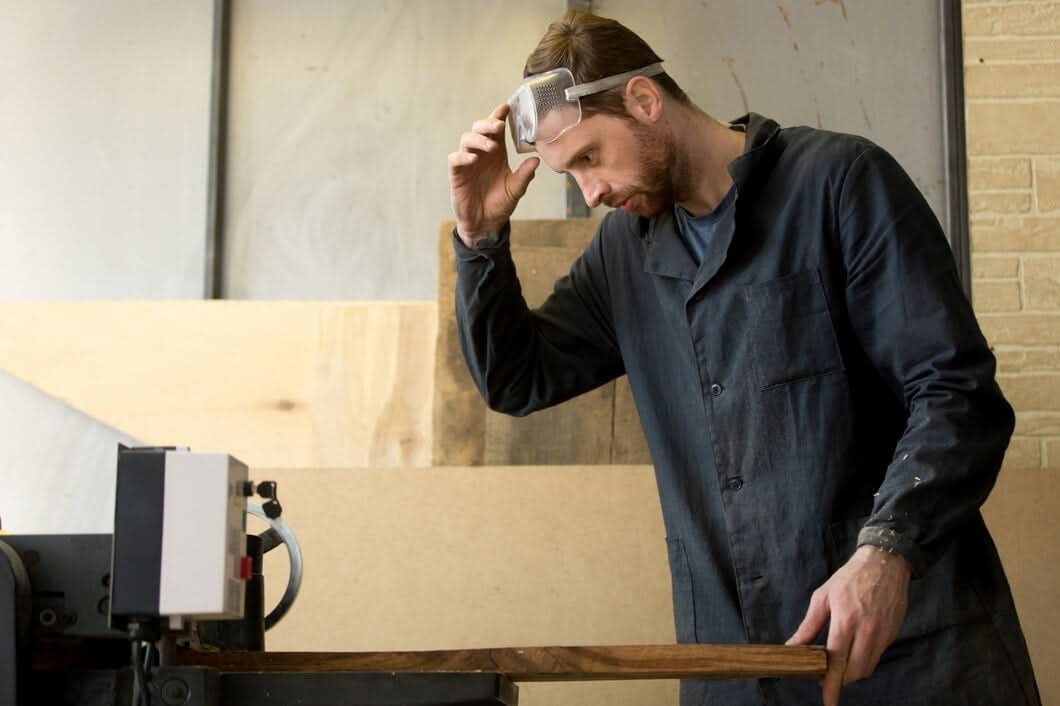 A professional carpenter wearing safety goggles meticulously operating a sawmill machine to cut a large slab of natural wood in a workshop.