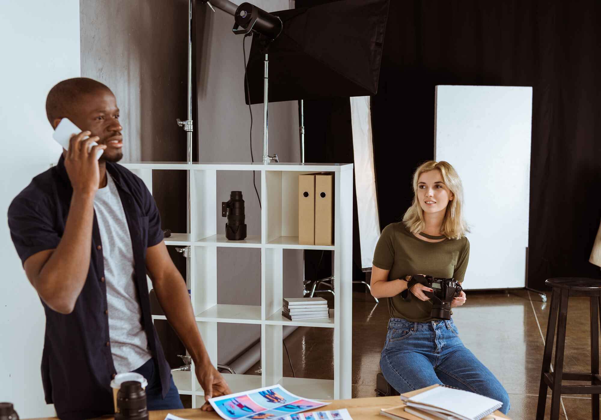 A male producer talking on a smartphone while a female photographer holds a camera in a professional studio with lighting equipment.