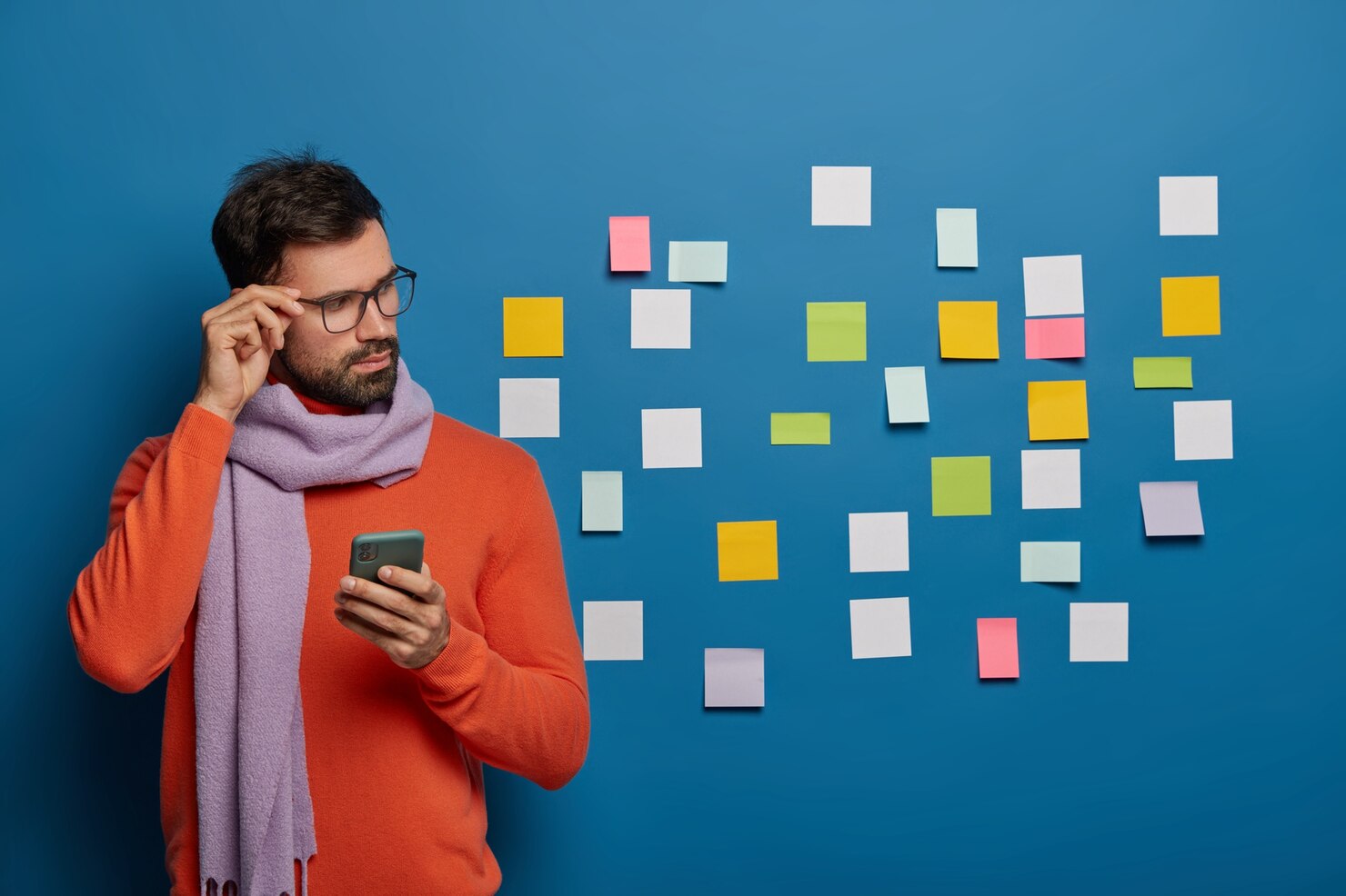 A stylish man wearing glasses and a scarf holding a smartphone next to a wall with sticky notes, representing digital marketing strategy in Boston, MA.