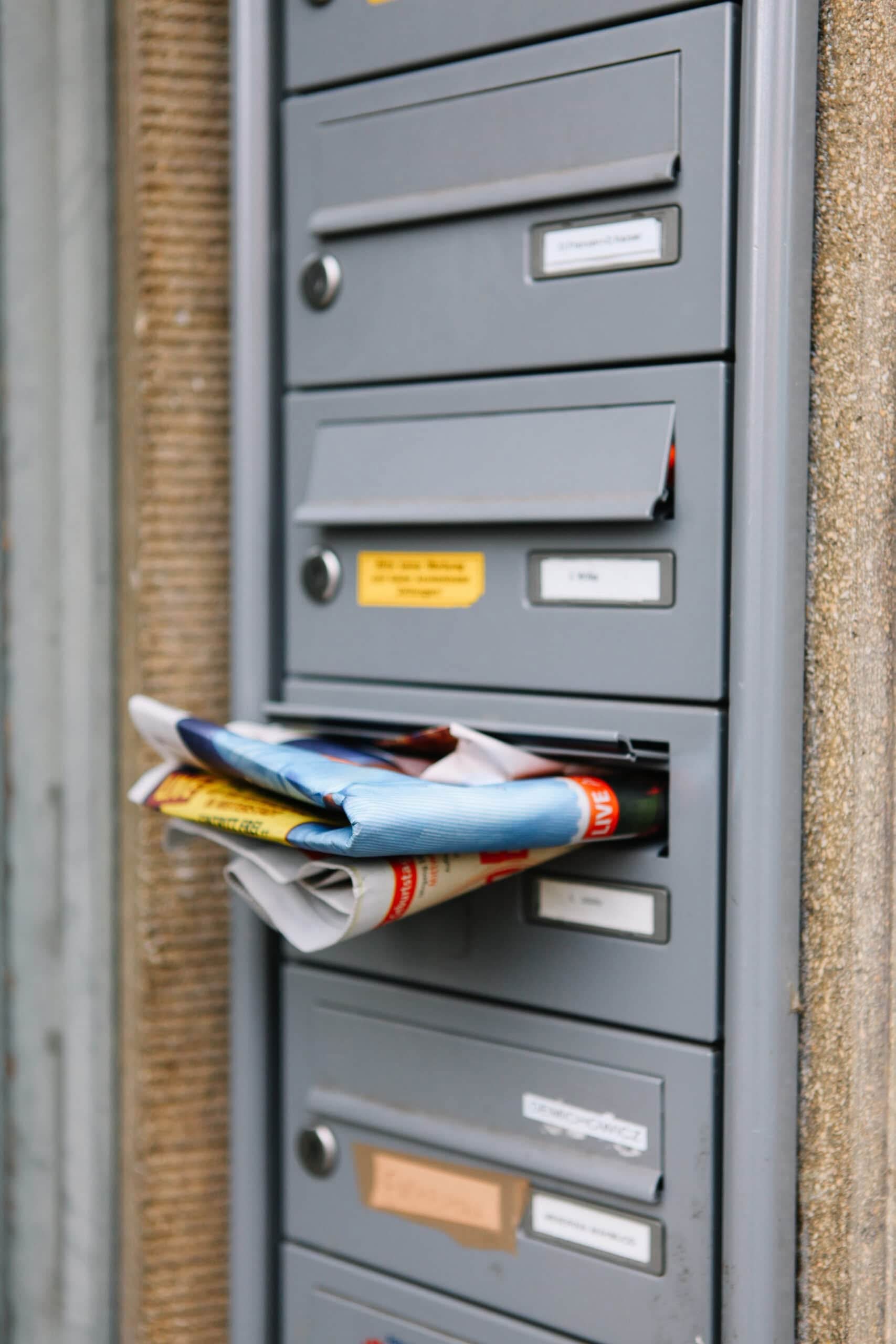 A vertical close-up of several gray apartment mailboxes, with one mailbox overflowing with folded newspapers and colorful direct mail flyers. Direct mail EDDM