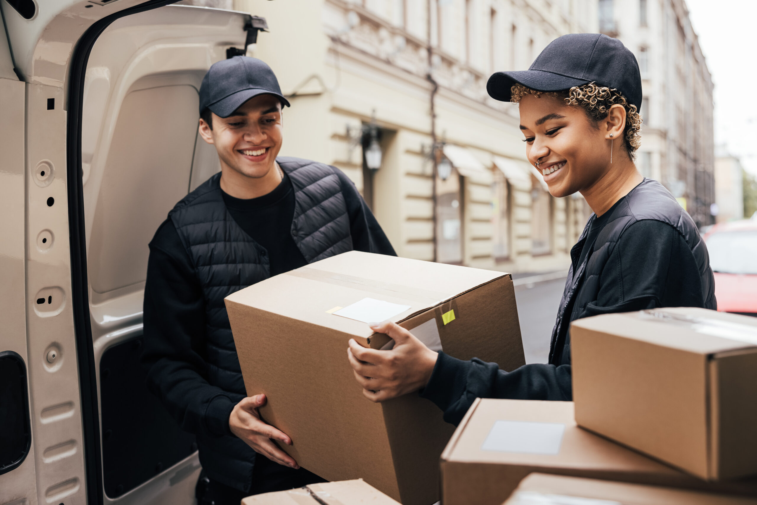 Two smiling couriers in black uniforms and caps unloading cardboard shipping boxes from the back of a white delivery van on a city street.