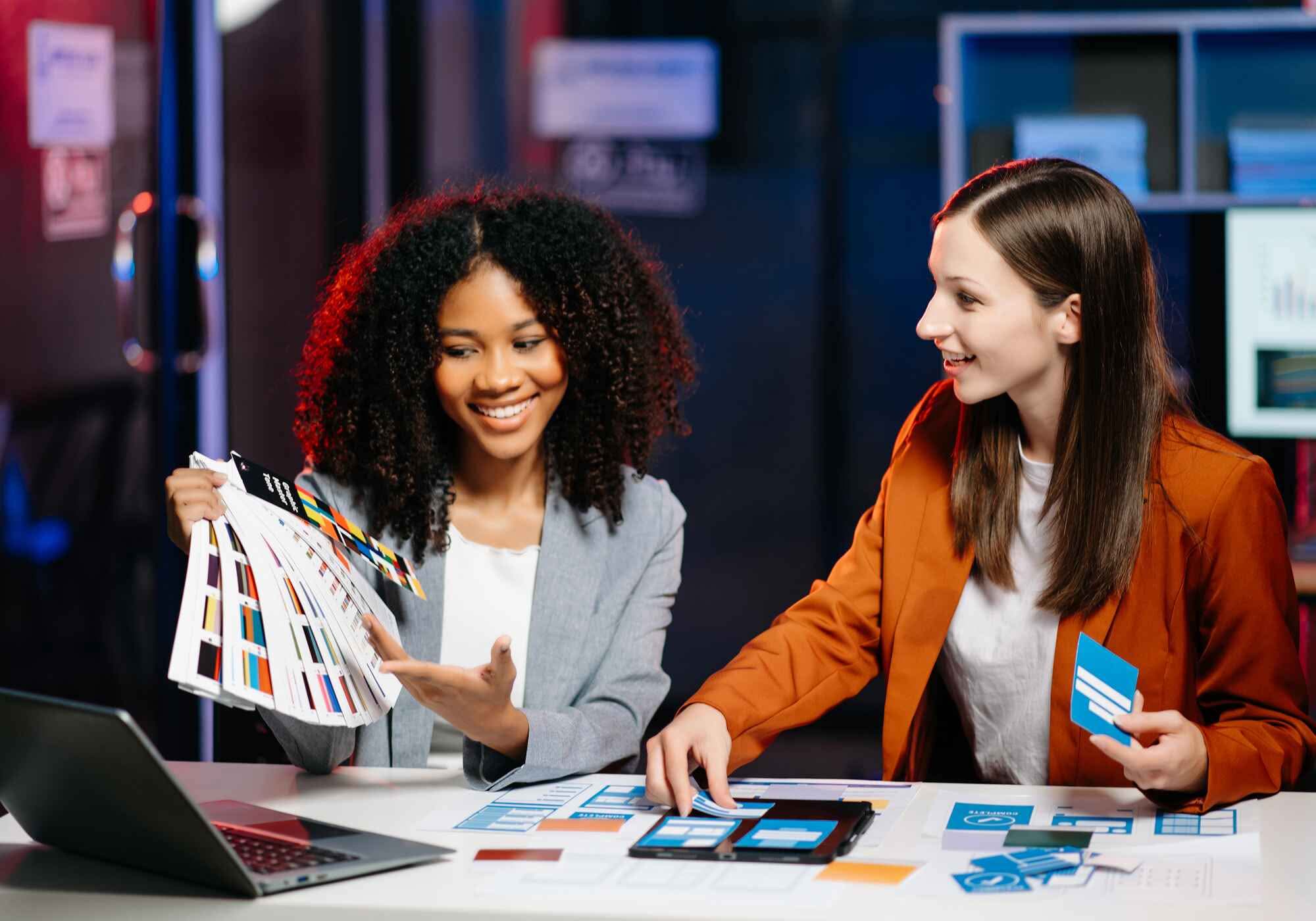 A UX developer and UI designer collaborate at a desk, reviewing mobile app wireframes and a color swatch fan in a modern office. Start A project from Brick City Branding