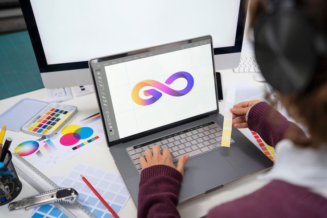 A graphic designer working on a laptop, creating a colorful 3D infinity loop logo, with color swatches and design tools visible on the desk.