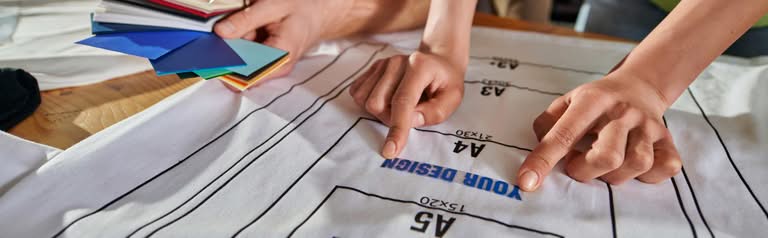 A close-up view of a designer's hands pointing at an A4 apparel print sizing template on a white garment while holding a vibrant color fabric swatch book for custom screen printing austin tx.